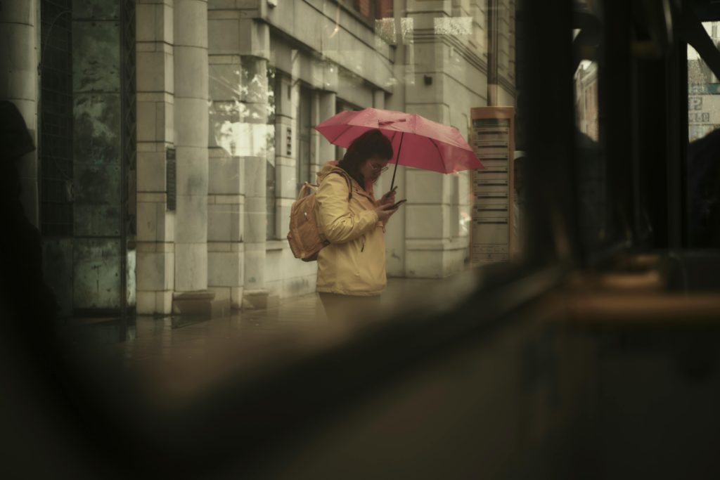 Não encontrou a vaga que deseja? 9 A woman with a pink umbrella checks her phone on a rainy city street, evoking a moody, candid feel.