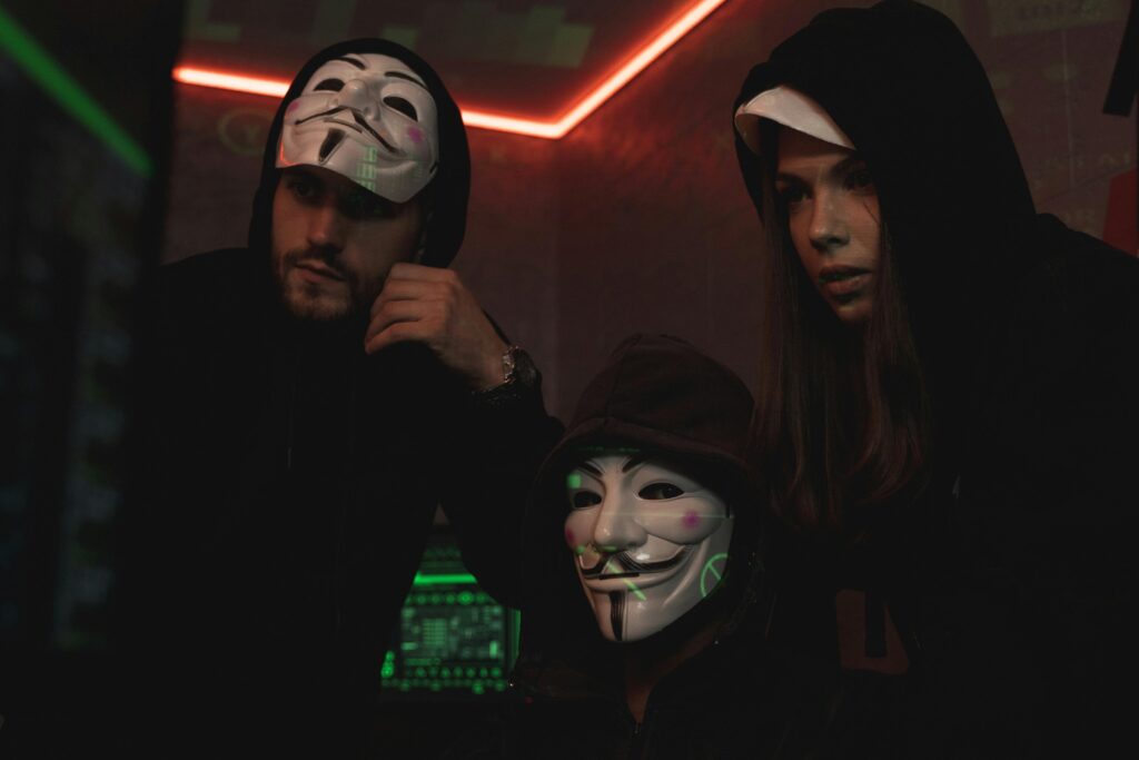 Golpes de emprego (Falsas vagas) 12 A group of people wearing Guy Fawkes masks in a dark tech-themed room, suggesting anonymity and hacking activities.